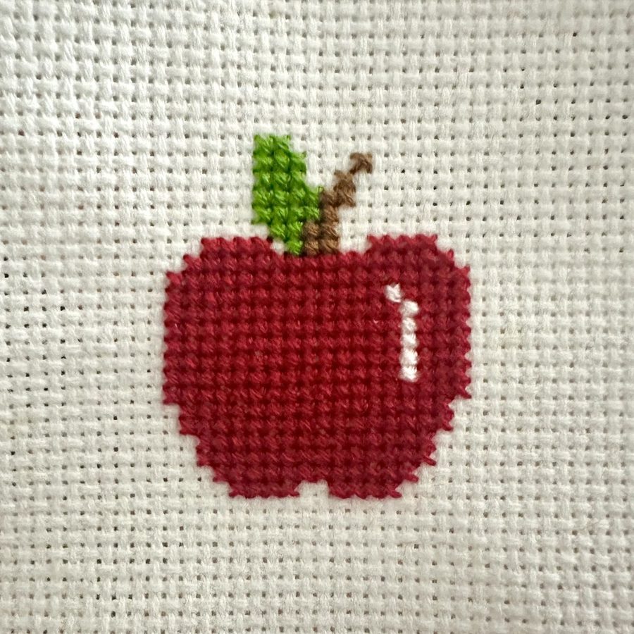 A small cross-stitched red apple with a green leaf, worked on white aida cloth